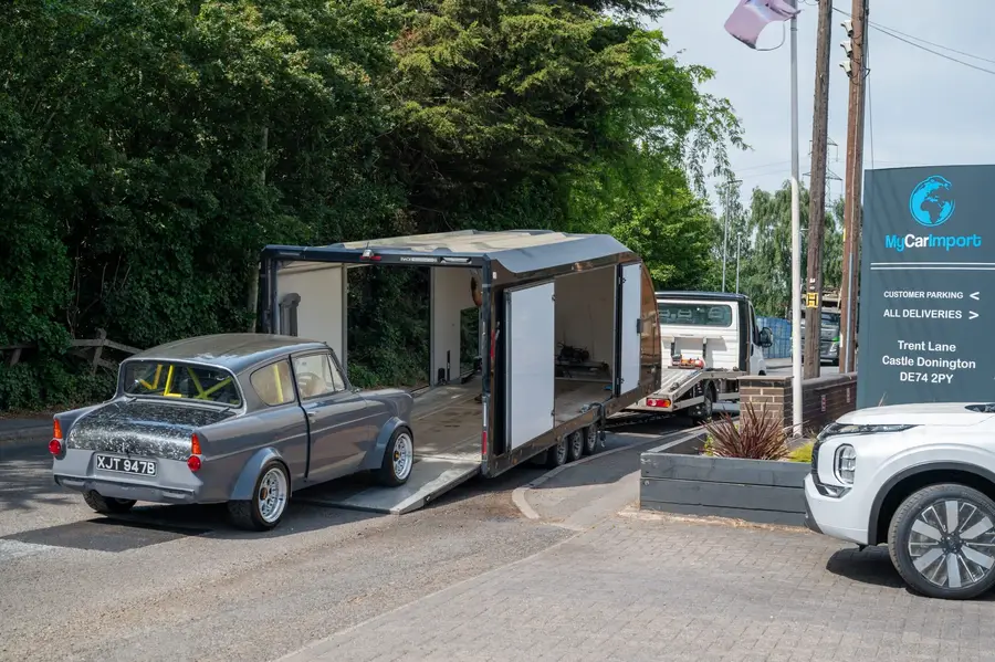 A grey Ford Anglia with a roll cage beside an enclosed car transporter, next to the My Car Import sign on Trent Lane, Castle Donington